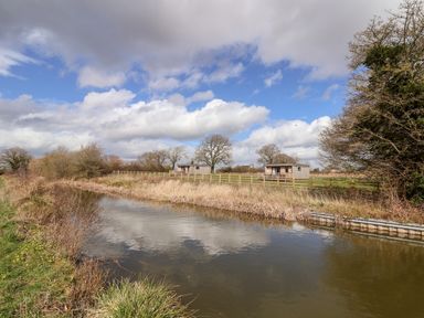 Two wooden cabins beside a fenced field with a river and trees under a cloudy sky at The Wren in Wem