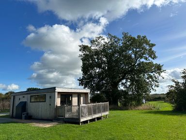 A small wooden cabin with a deck on green grass under a partly cloudy sky at The Wren in Wem