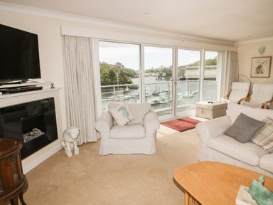 A living room with white armchairs and a sofa facing a glass door with a view of boats on water at Creekside House in Fowey