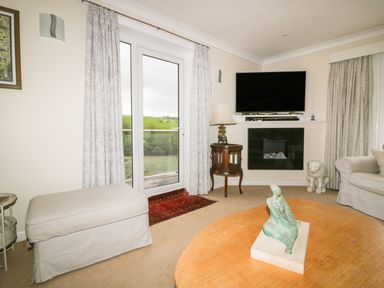 A living room with a beige ottoman wooden side table with lamp flat screen tv wall fireplace statue on a wooden coffee table and glass doors at Creekside House in Fowey