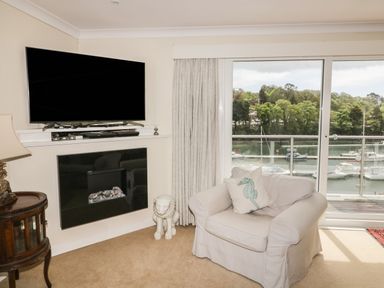A living room with a large armchair a seahorse cushion a wall-mounted television and glass doors overlooking boats on water at Creekside House in Fowey