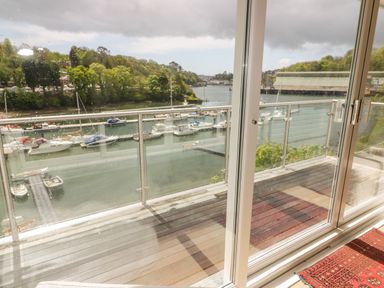 A balcony with wooden flooring and glass railing overlooking boats docked on a river and green trees beyond at Creekside House in Fowey