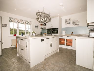 A kitchen with a central island, hanging pots and pans, large window, grey floor, and white cabinets at Creekside House in Fowey