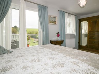 A bedroom with a bed a wooden nightstand a wooden wardrobe and windows with blue curtains at Creekside House in Fowey