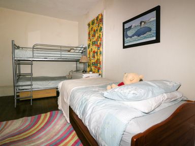 A bedroom with single bed and bunk beds a nightstand with lamp colorful curtains and a framed whale picture at Creekside House in Fowey