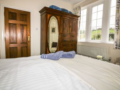 A bedroom with a wooden wardrobe with mirror a wooden door folded towels on a bed and windows with curtains at Creekside House in Fowey