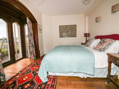 A bedroom with a bed covered in a blue quilt and floral pillows near wooden framed windows and floral curtains at Creekside House in Fowey