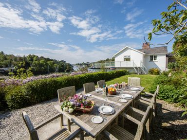 An outdoor dining table with plates and drinks in a garden with a white house and water view at Creekside House in Fowey