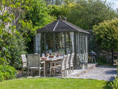 A garden patio with a wooden dining table and chairs next to a glass gazebo and a barbecue grill at Creekside House in Fowey