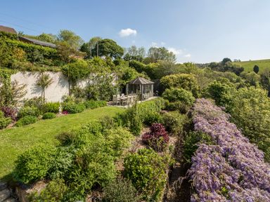 A garden with a patio dining set and a gazebo surrounded by shrubs and flowering plants at Creekside House in Fowey