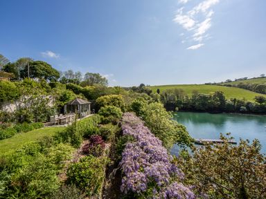 A garden with a dining table and chairs beside a small building overlooking a river at Creekside House in Fowey