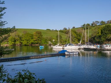 A small marina with sailboats and motorboats docked on a calm river with green hills and trees in the background at Creekside House in Fowey