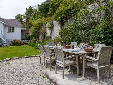 An outdoor dining area with a wooden table and chairs set on gravel near a garden and white house at Creekside House in Fowey