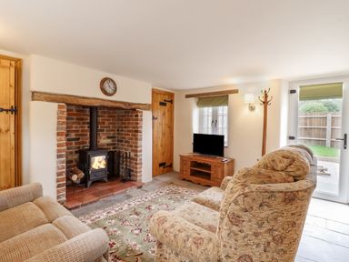 A living room with a brick fireplace a wood stove two upholstered armchairs a wooden TV stand with a television a coat rack and a door leading to a garden at Postbox Cottage in Feltwell
