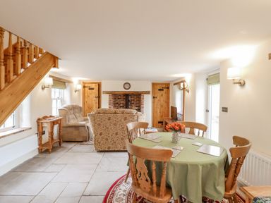A dining area with a round table and chairs next to a sitting area with upholstered chairs and a brick fireplace at Postbox Cottage in Feltwell