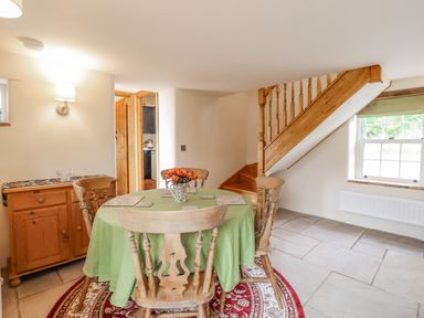 A dining area with a round table covered with a green cloth surrounded by wooden chairs next to a wooden staircase at Postbox Cottage in Feltwell