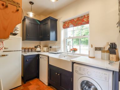 A kitchen with dark cabinets, a white farmhouse sink, a washing machine, a dishwasher, a refrigerator, and a window with a red patterned blind at Postbox Cottage in Feltwell