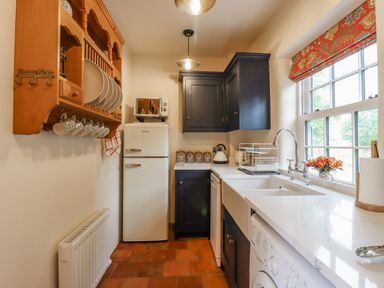 A kitchen with a white refrigerator wooden shelf with plates and mugs dark cabinets and a window with floral blind at Postbox Cottage in Feltwell