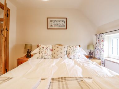 A bedroom with a double bed with floral and animal print pillows two bedside tables with lamps a window with floral curtains and a framed picture above the bed at Postbox Cottage in Feltwell