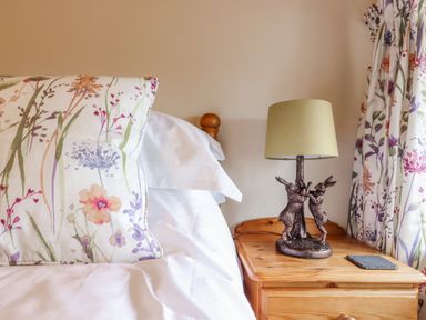 A bedroom with a floral pillow on a bed and a wooden nightstand with a rabbit lamp at Postbox Cottage in Feltwell