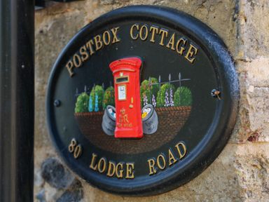 A decorative black circular plaque with a red postbox and garden scene showing Postbox Cottage 80 Lodge Road