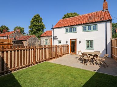 A backyard with a wooden fence a patio with a round table and four chairs and a white house with a red tiled roof at Postbox Cottage in Feltwell