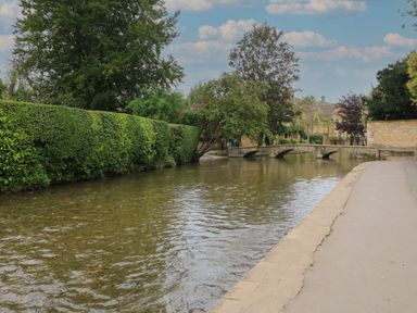 A shallow river with a stone bridge and trimmed hedges beside a paved walkway at Tuesday Cottage in Bourton-On-The-Water