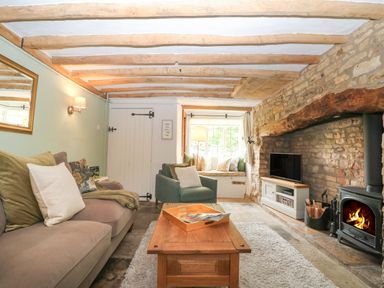 A living room with wooden beams on ceiling a stone fireplace with a wood stove a sofa an armchair and a window seat at Tuesday Cottage in Bourton-On-The-Water