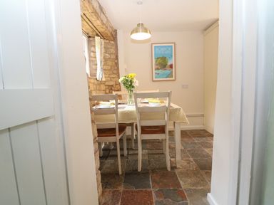 A dining area with a table covered by a yellow and white tablecloth four chairs a vase with flowers and a framed wall poster at Tuesday Cottage in Bourton-On-The-Water
