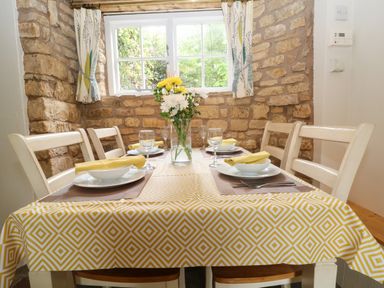 A dining table set for four with plates glasses napkins and flowers by a window at Tuesday Cottage in Bourton-On-The-Water