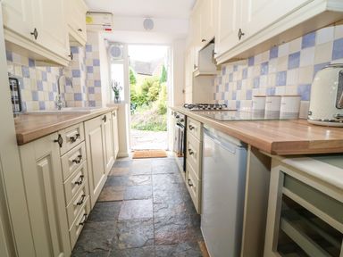 A narrow kitchen with wooden countertops white cabinets blue and white tiled backsplash and stone floor at Tuesday Cottage in Bourton-On-The-Water