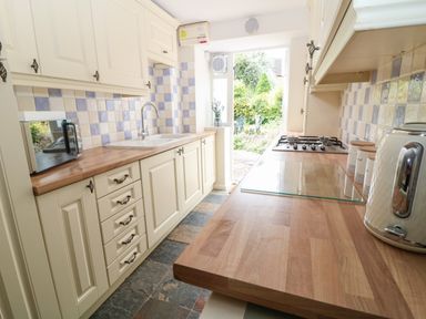 A galley kitchen with wooden countertops cream cabinets a gas stove a microwave toaster and an open door leading to a garden at Tuesday Cottage Bourton-On-The-Water