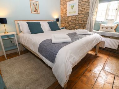 A bedroom with a bed with white and gray bedding two nightstands with lamps a stone wall and a window seat with cushions at Tuesday Cottage in Bourton-On-The-Water
