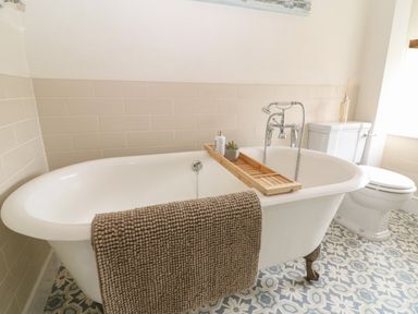 A bathroom with a clawfoot bathtub with a towel and bath tray next to a toilet with patterned floor tiles at Tuesday Cottage in Bourton-On-The-Water