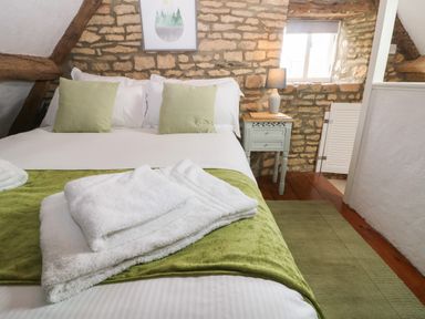 A bedroom with a bed white linens green cushions and blanket a bedside table with a lamp and a stone wall at Tuesday Cottage in Bourton-On-The-Water