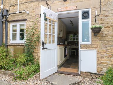 A stone cottage exterior with an open white door showing a kitchen inside at Tuesday Cottage in Bourton-On-The-Water