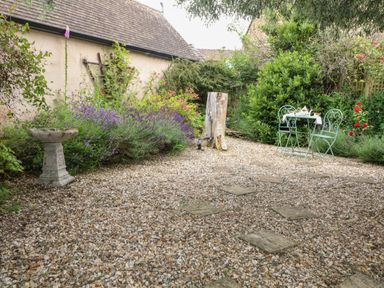 A garden with gravel ground two metal chairs and a table with tea set surrounded by plants and flowers at Tuesday Cottage in Bourton-On-The-Water