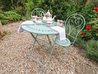 An outdoor metal table with two chairs tea set and white napkins on gravel surrounded by plants and flowers at Tuesday Cottage in Bourton-On-The-Water