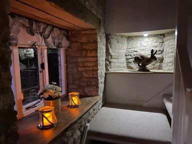 A stone walled stair landing with two lit candles and a flower pot on the windowsill and a rabbit sculpture in a niche at Tuesday Cottage in Bourton-On-The-Water