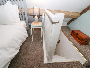 A bedroom corner with a bed white bedding a small side table with a lamp a wooden chest on a ledge and a staircase with white railing