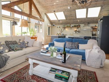 A living room with two sofas a wooden coffee table with books flowers and a bottle of wine and a kitchen in the background at The Owl's Shack in Kendal