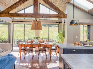 A dining area with a wooden table and chairs next to large windows and a kitchen island with items on the counter at The Owl's Shack in Kendal
