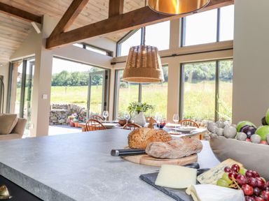 A kitchen island with bread cheese and fruit with dining table and chairs near large windows at The Owl's Shack in Kendal