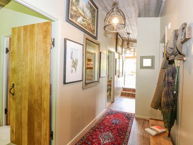 A hallway with framed pictures on the wall a wooden bench with coats and hats a red patterned rug and hanging lights at The Owl's Shack in Kendal