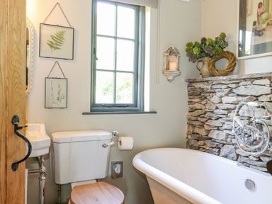 A bathroom with a white bathtub a toilet with a wooden seat a small sink stone wall and window at The Owl's Shack in Kendal