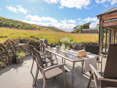 An outdoor patio with a glass table and chairs overlooking a grassy field at The Owl's Shack in Kendal