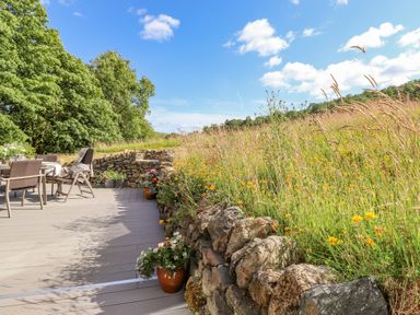 An outdoor patio with chairs and table next to a stone wall and wildflowers at The Owl's Shack in Kendal