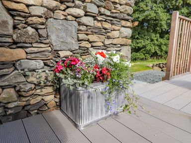A metal planter with red white and purple flowers against a stone wall on a wooden deck at The Owl's Shack in Kendal
