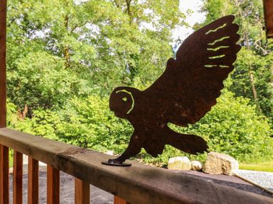 A metal owl silhouette decoration on a wooden railing with trees and rocks in the background at The Owl's Shack in Kendal