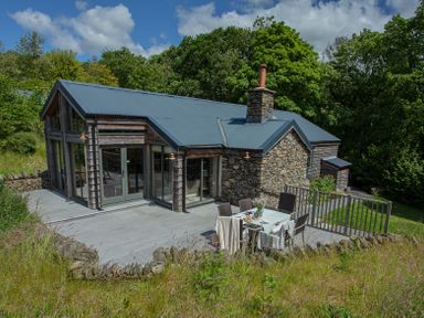 A stone and wood house with a metal roof and outdoor dining table on a patio surrounded by trees at The Owl's Shack in Kendal